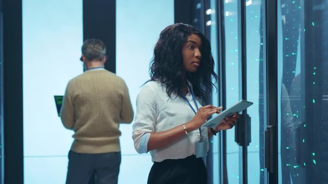 Beautiful afro young woman IT specialist visually inspecting modern secure server racks cabinet coworking with man colleague inside professional data center.
