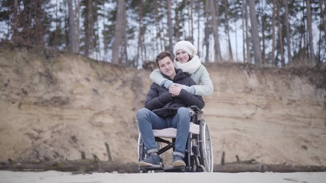 Positive handsome Caucasian boy in wheelchair and beautiful adult woman looking at camera and smiling. Cheerful disabled young man enjoying time outdoors with mother or invalid tender.
