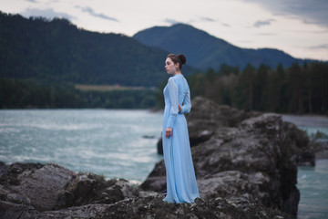 A girl in a blue dress stands on the banks of the river.