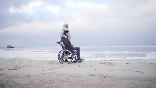 Side view of young Caucasian woman and boy in wheelchair standing on sandy river bank. Wide shot portrait of mother and son or invalid tender and patient enjoying autumn day outdoors.