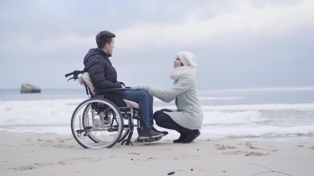 Young woman sitting on hunkers in front of boy in wheelchair and talking. Mother or invalid tender supporting handicapped young man on sea shore or river bank outdoors. Joy, lifestyle, help.
