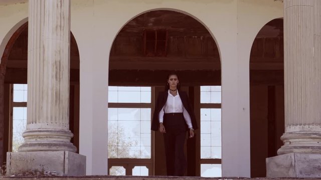 fashionable young woman in pantsuit and white shirt goes from the classic abandoned building to the terrace with two columns