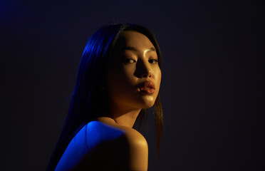 Profile of a young beautiful Asian girl, she looks directly at the camera. The hair and the back of the woman in bright , bright blue ultraviolet lights. On black background.