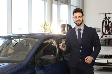 Young salesman with key and clipboard near car in dealership