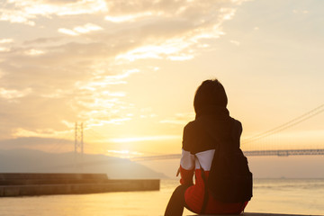 Silhouette alone young woman relax siting on riverside ocean and watching sunset and cloud sky with akashi kaikyo bridge. Thinking and concentrate in summer background with copy space for label text.