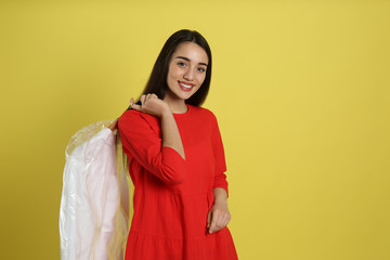 Young woman holding hanger with shirt in plastic bag on yellow background. Dry-cleaning service