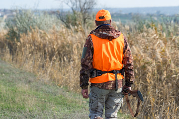 A man with a gun in his hands and an orange vest on a pheasant hunt in a wooded area in cloudy weather. Hunter with dogs in search of game.