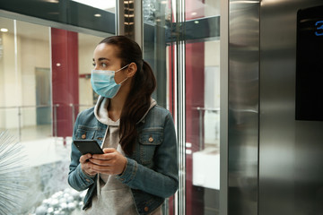 Young woman with disposable mask and smartphone in elevator. Dangerous virus