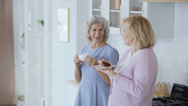 Medium shot of two senior female friends standing in domestic kitchen, having tea and cake and chatting