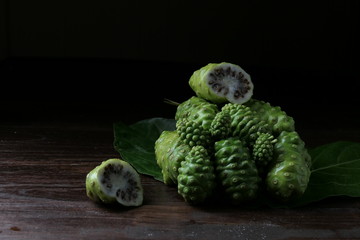 Noni fruit, herbs and sliced on wood, on a black background. Dark tones, rich in nutrients. Antioxidants.