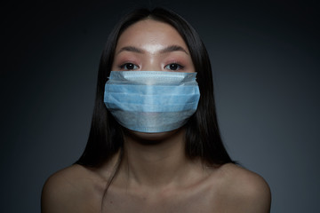    A young beautiful Asian woman in a medical mask, on an isolated gray background, looks directly...
