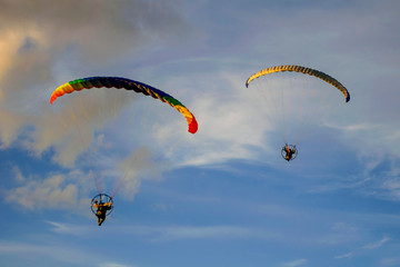 Two brightly colored paragliders fly in the blue sky with white clouds.