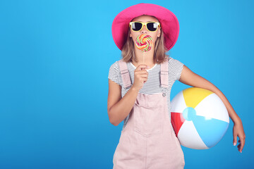Young girl wearing sunglasses, hat and holding lollipop and inflatable ball on blue background