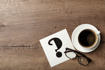Note with question mark, eyeglasses and cup of coffee on wooden table, flat lay. Space for text