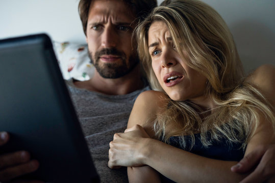 Young couple watching horror movie on digital tablet in bedroom