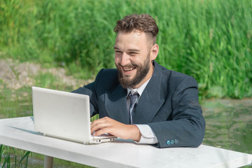 A smiling bearded man in a business suit sits at a white table in a swamp and looks at a laptop. manager performs office work