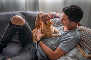 joven tumbado en un sofá debajo de la ventana, abraza y acaricia a un pequeño gato atigrado