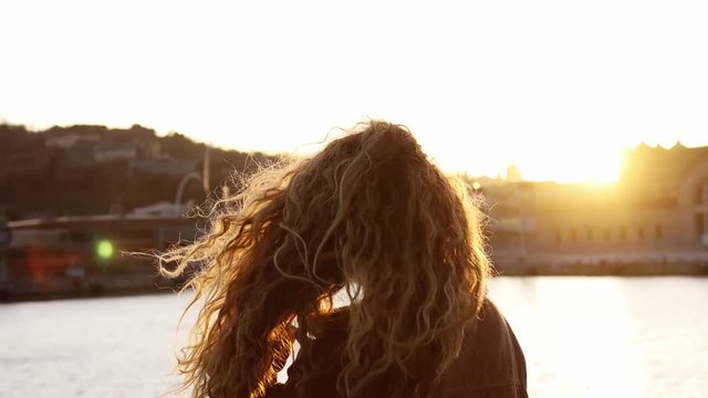 Blond girl with curly hair watching the sunset in the golden hour. Wind mooving her hair.