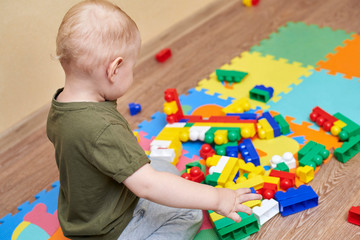 A two-year-old boy plays with a colorful constructor set on the floor of a house.