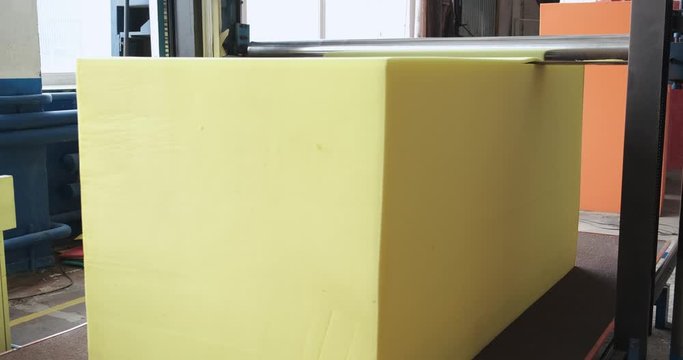 Large yellow cube of synthetic foam is on machine conveyor, rotating knife cuts foam into thin layers, premises of factory floor, precise cutting of raw materials, semi-automatic, foam rubber products