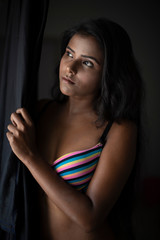 Portrait of young and beautiful dark skinned Indian Bengali woman in colorful lingerie/bikini and hot pants standing in front of window and looking outside in studio background. Boudoir photography.
