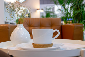 A cup of coffee or tea on a small table in a light cafe with green plants and leather armchairs. White vase with white flowers on the table. Blurred background. Selective focus. Horizontal orientation