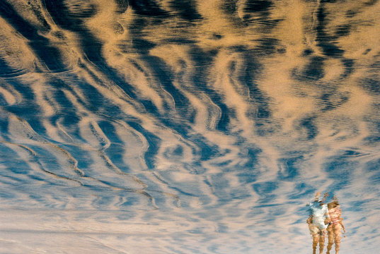 Reflection in the sand of a couple walking on the beach.