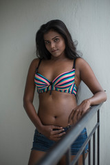 Portrait of an young and beautiful dark skinned Indian Bengali woman in colorful lingerie/bikini and hot pants posing in casual mood on a balcony in white urban background. Boudoir photography.