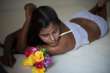 Obraz na płótnie Canvas Portrait of an young dark skinned Indian Bengali woman in lingerie and vibrant flowers lying on white bed in casual mood in white background. Indian lifestyle and boudoir photography.
