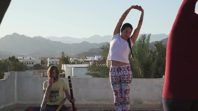 Teacher leading yoga group on rooftop while man plays didgeridoo instrument