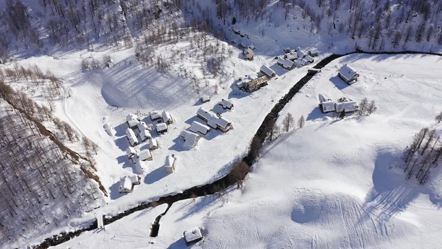 aerial over winter wonderland, snowy village in sunny winter day.Winterland scene with lodges and cottages near small creek.Mountains outdoor establisher.4k drone forward flight establishing shot