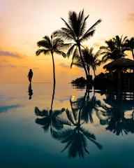 Silhouette of Beautiful Woman Walking on Edge of Infinity Pool in Vacation Paradise of Hawaii with Tropical Island Background of Palm Trees and Colorful Sunset Sky at Dusk with Reflection in Water