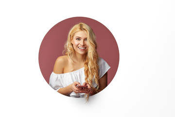 Image of young happy woman standing over white background. Girl peep out of a hole in the wall. Looking to the side to take a selfie on the mobile phone.