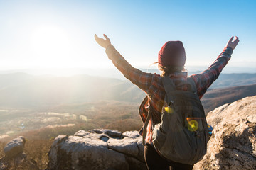 Young Woman Hiking on Mountain Top With Arms Raised Wearing Backpack and Beanie Looking Out at Colorful Landscape Horizon of Mountain Range with Bright Blue Sky and Sunshine Sun Rays