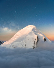 Beautiful Aerial Nature Photo of Winter Scene with Snow Covered Mountain Peak and Colorful Sunset Light with Amazing Night Sky Background Orange and Blue with Stars and Milky Way Galaxy Above Clouds