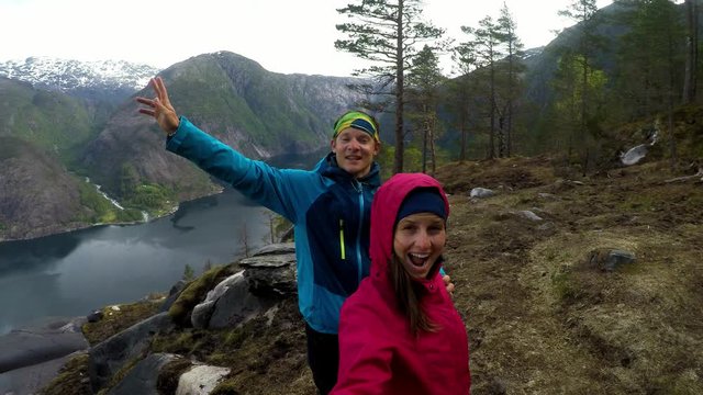 Couple standing next to Langfossen Waterfall and amazing fjord view on a cloudy day in Norway. The other side of the fjord is partially covered with snow. Navy blue water contrasted with green slopes
