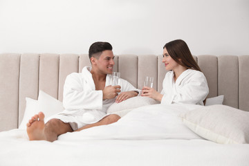Happy couple in bathrobes with glasses of water on bed at home