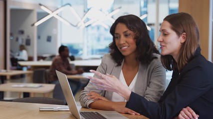 Two businesswomen with laptop at desk in open plan office collaborating on project together - shot in slow motion - Powered by Adobe