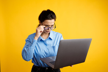 Serious businesswoman in glasses holding laptop and using it isolated over the yellow background