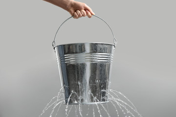 Woman holding leaky bucket with water on light grey background, closeup - obrazy, fototapety, plakaty