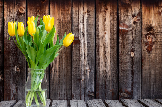 Bunch of bright yellow spring tulips in a glass container on a rustic plank table.