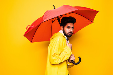 Young handsome bearded indian man in yellow raincoat with red umbrella cover from rain isolated over orange background