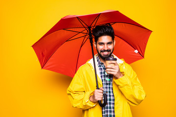 Young handsome bearded indian man in yellow raincoat with red umbrella cover from rain holding coffee to go isolated over orange background