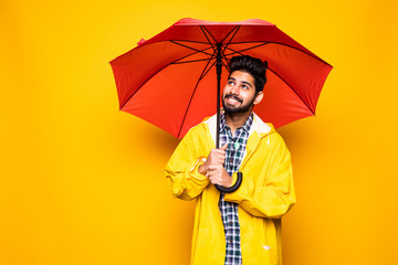 Young handsome bearded indian man in yellow raincoat with red umbrella cover from rain isolated...