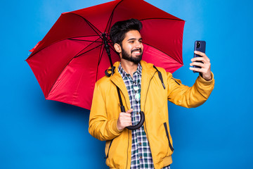 Young handsome bearded indian man in yellow raincoat with red umbrella cover from rain use phone isolated over blue background