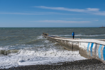 Winter sea landscape. A girl in a blue jacket stands on a breakwater and looks at the waves.