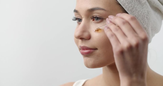 Side View Of Attractive Woman Applying Cosmetic Oil From Pipette On Her Clean Face Over White Studio Background. Concept Of Facial Treatment And Moisturizing