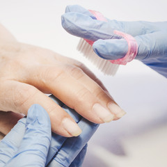 Manicure beauty salon master in blue rubber gloves uses brush to remove dust from client nails on white background
