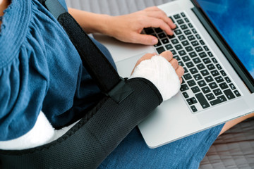 Freelancer woman with broken arm in cast and bandage sits on bed in medical hospital after the manipulations of medical worker and works on laptop. Accident. Orthopedic problem. Lifestylе