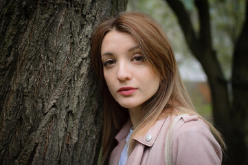 Naklejka premium A young attractive woman with a blonde hair posing near a big tree in a spring park outdoors. She leans against a tree to touch the bark